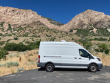 White Ford Transit rv rental camper van, equipped with an AC unit, parked on asphalt before rocky mountains and clear sky.
