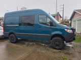 Teal adventure van, an ideal RV rental, parked on a wet drive. Features include off-road tires and roof solar panels.