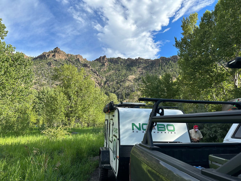 A NO BO travel trailer (RV rental option) is hitched to a truck, parked in a lush mountain landscape. Man in red hat seen. — photo 18