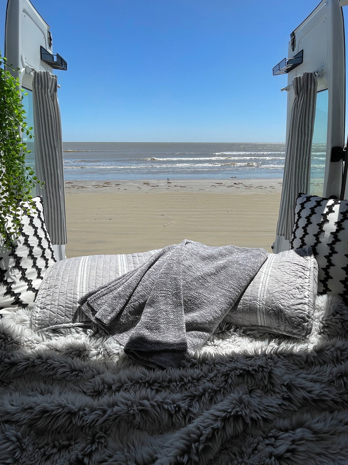 Perfect RV rental view: sunny beach & ocean from a cozy interior. Pillows, blankets, and faux fur invite relaxation.