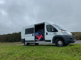 Person in a serene pose inside a white RV rental van, parked on a grassy hill beside a road under an overcast sky.