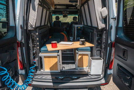 RV rental camper van's open rear reveals a mobile living space with a table, seating, utility panel, and a coiled blue hose.