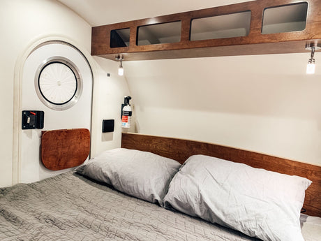 Comfortable RV rental bedroom with a cozy bed, wooden overhead storage, and a unique circular door in a compact camper interior.