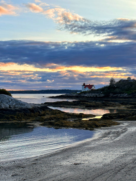 Coastal beauty at sunset! A lighthouse on a rocky shore, with calm water reflecting the colorful sky. Perfect for an RV rental trip.