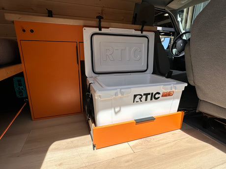 Inside an RV rental: custom orange storage, a sliding RTIC cooler, and light wood-look flooring for adventure.