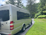 A silver Mercedes-Benz Weekender RV rental is parked on a shaded driveway with lush green grass and tall trees.