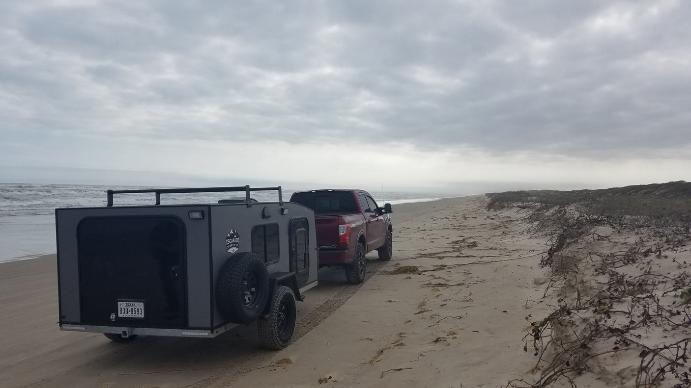 Red pickup tows grey RV rental on sandy beach by ocean, ideal for off-road camping and coastal adventures.