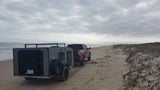Red pickup tows grey RV rental on sandy beach by ocean, ideal for off-road camping and coastal adventures.