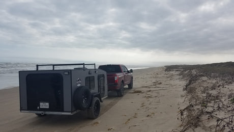 Red pickup tows grey RV rental on sandy beach by ocean, ideal for off-road camping and coastal adventures.