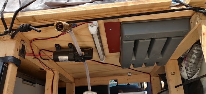 Underneath an RV rental setup: a 12V water pump, clear tubing, wiring, LED lights, and storage optimizing mobile living.