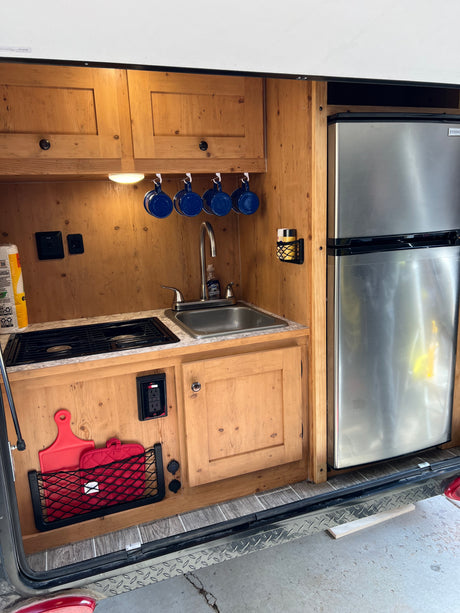 Experience mobile living! This RV rental features a compact kitchen with a sink, stove, fridge, and wooden cabinetry.
