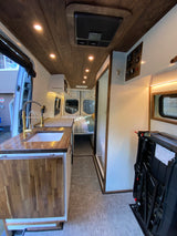 A modern RV rental interior: featuring a well-appointed kitchen with gold fixtures, wood ceiling, bed, and cozy living space.