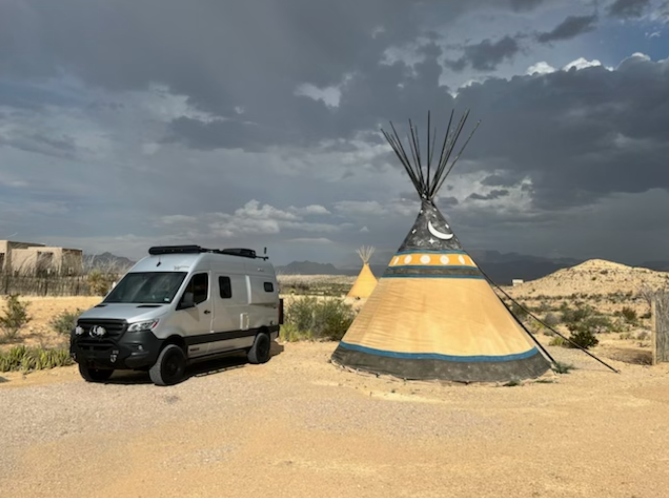Experience desert glamping! Silver RV rental parked near artistic teepees under dramatic clouds.