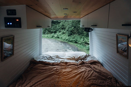 Inside an RV rental, a comfy bed looks out to a forest road. White paneled walls, wood ceiling, controls visible.