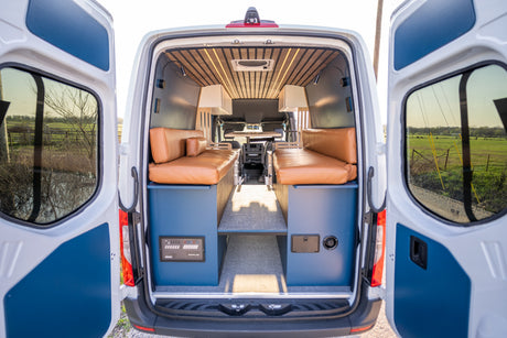 Inside a stylish RV rental: tan benches, wood ceiling, and back doors open to a tranquil green field and blue sky.