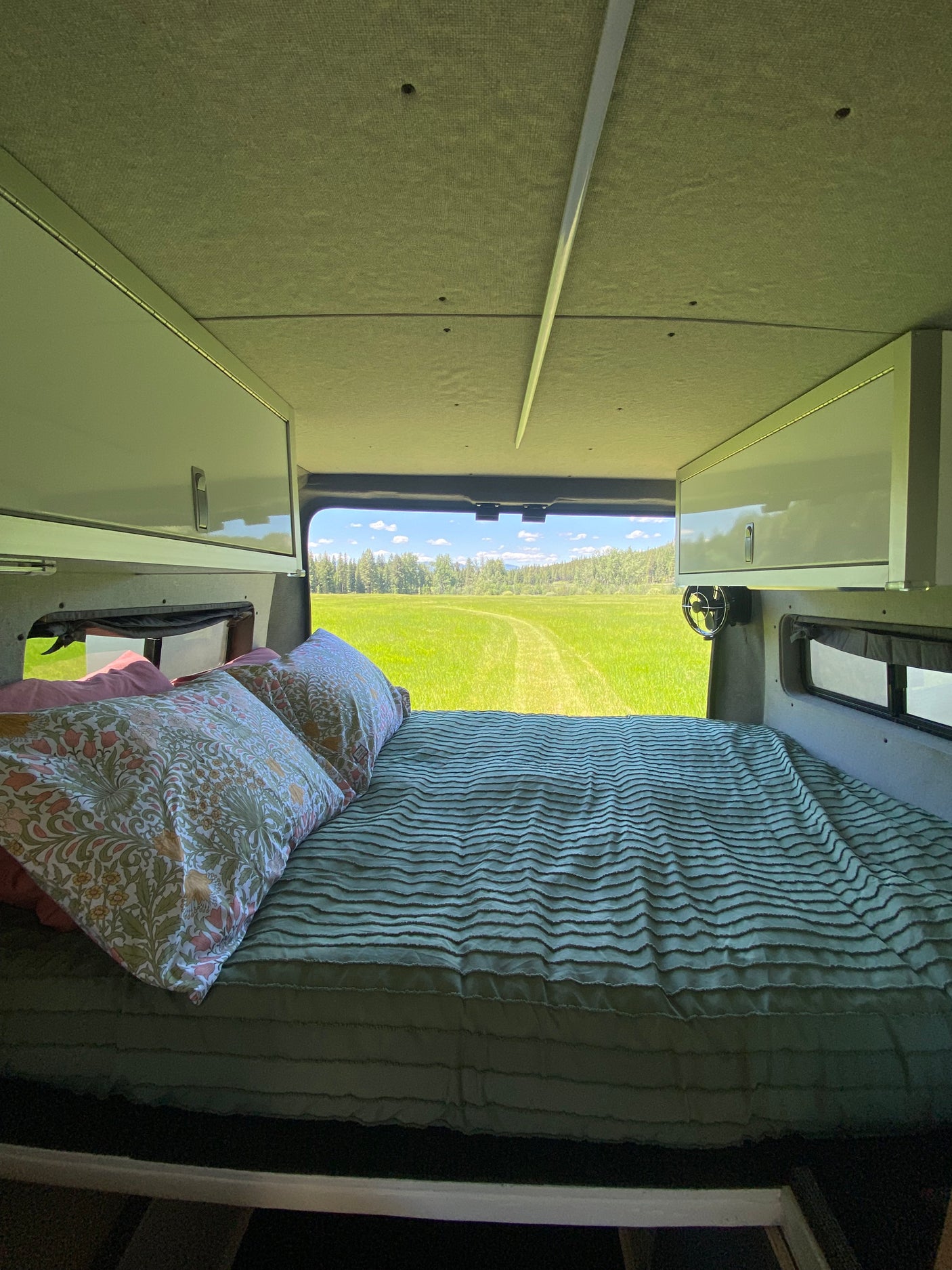 Inside a comfy RV rental: A bed with patterned pillows and green comforter offers a picturesque view of a lush green field and forest.