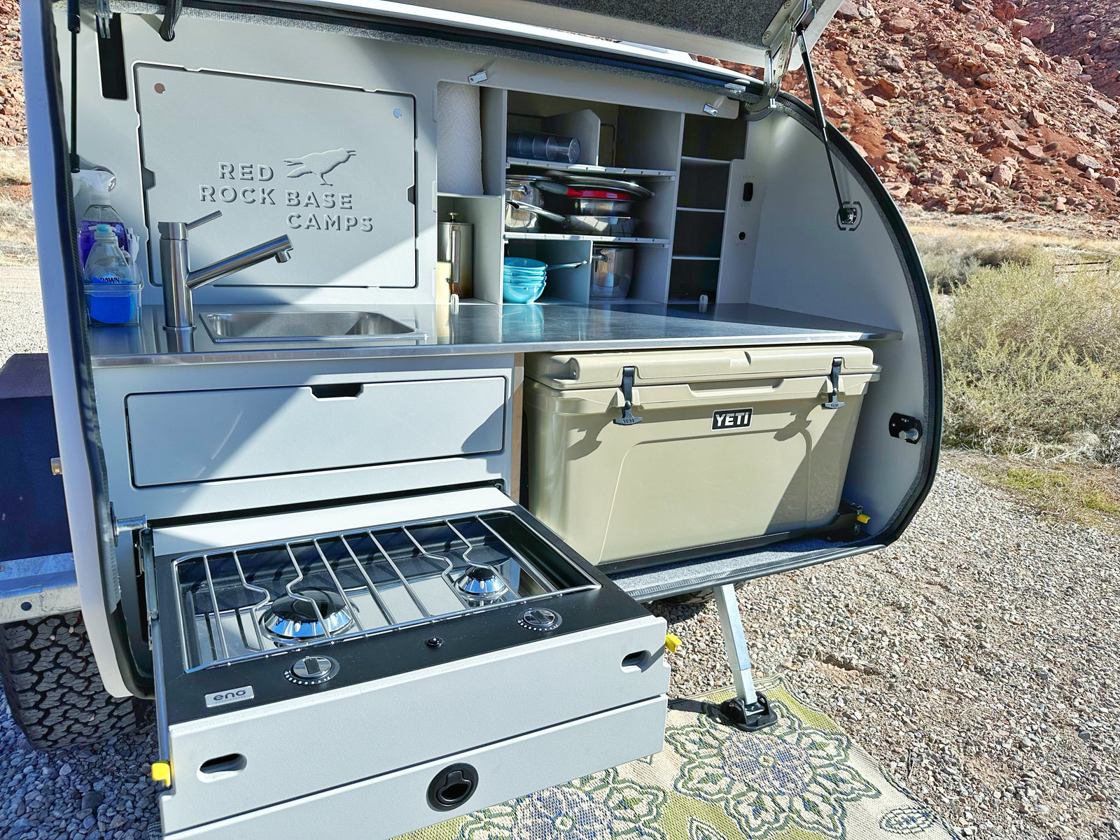 An RV rental showing a fully equipped pull-out outdoor kitchen with a sink, stove, and cooler, ideal for camping.