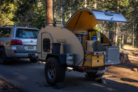 Overland Equipment teardrop RV rental setup with outdoor kitchen, ready for camping in a forest. Lexus SUV attached.