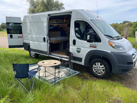 This white Ram ProMaster camper van, available for RV rental, offers an outdoor camping setup with chairs, table, and rug.