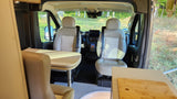 Inside an RV rental, showing beige swivel front seats, a dining table, and counter space, with a view of trees outside.