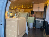 Inside a custom RV rental: kitchenette with sink, bed, and overhead storage, bathed in warm string lights.
