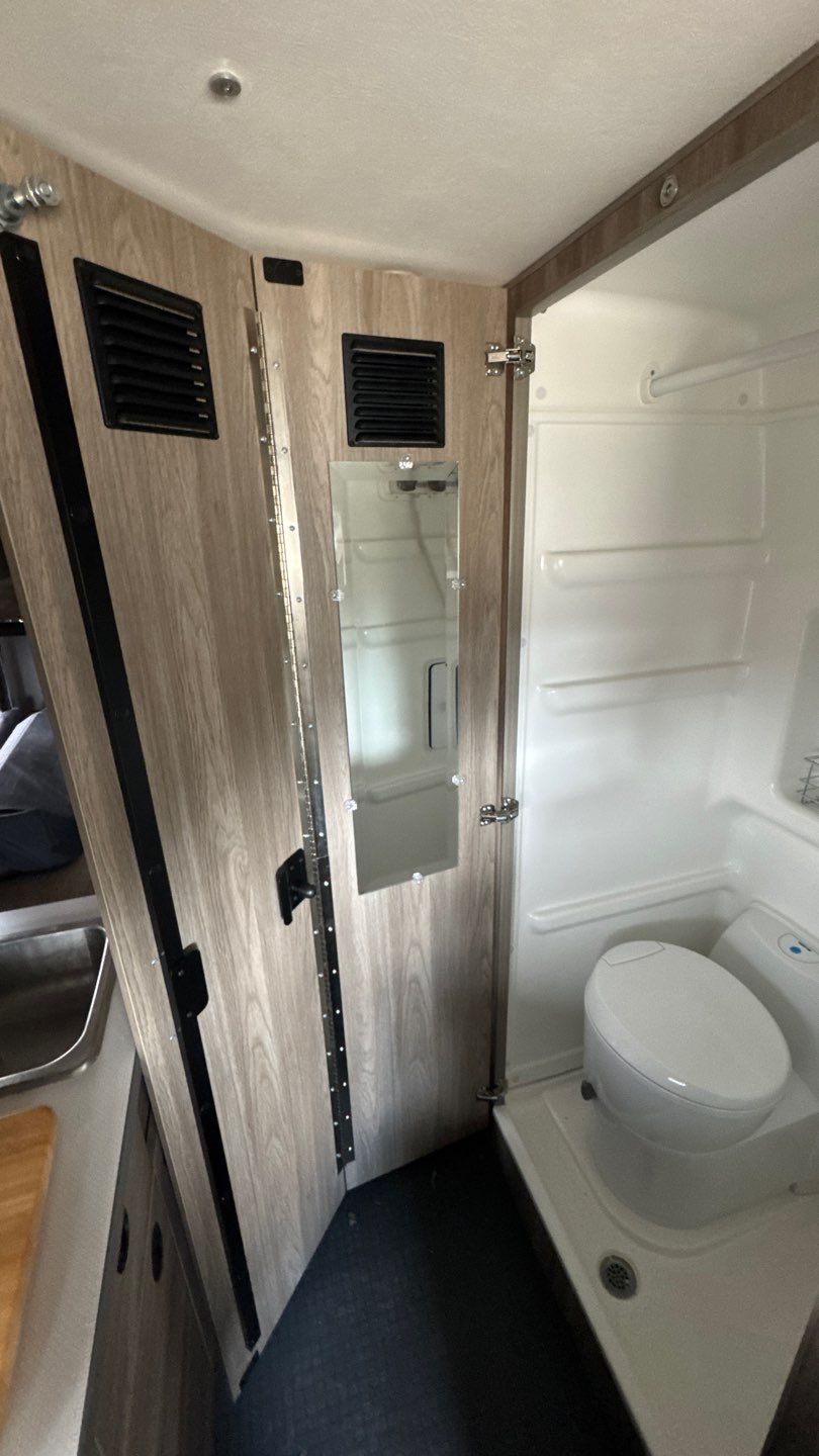 Compact rv rental wet bath: white toilet, shower basin, and adjacent sink. Light wood-grain walls, vents, and mirror.