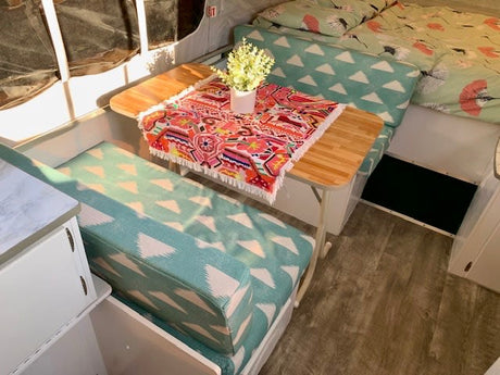 Cozy rv rental interior: bright dining area with teal patterned cushions, wooden table, colorful throw, and a comfy bed.