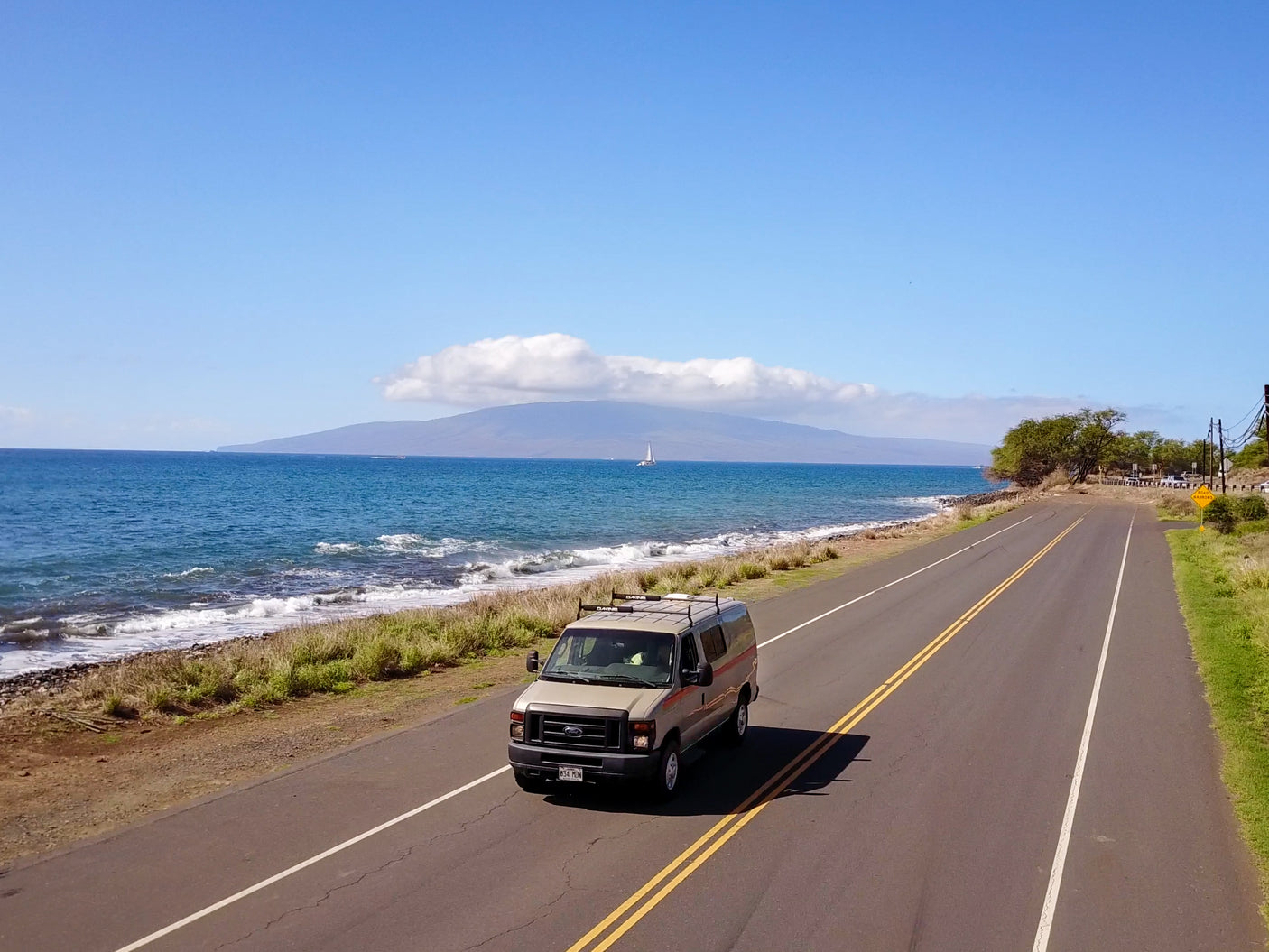 Scenic Hawaii coastal drive with an RV rental van and ocean views, sailboat in the distance.
