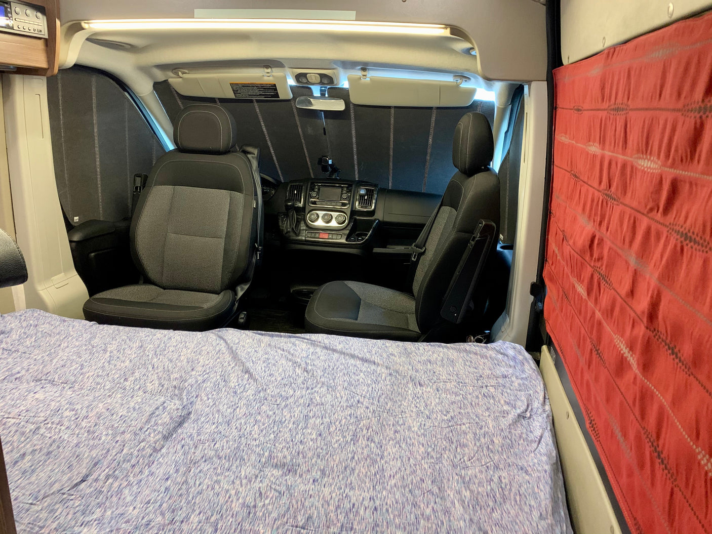 Interior of an RV rental: comfy bed in foreground, front cabin with driver/passenger seats & privacy screens visible.