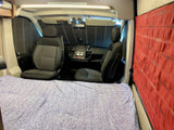 Interior of an RV rental: comfy bed in foreground, front cabin with driver/passenger seats & privacy screens visible.
