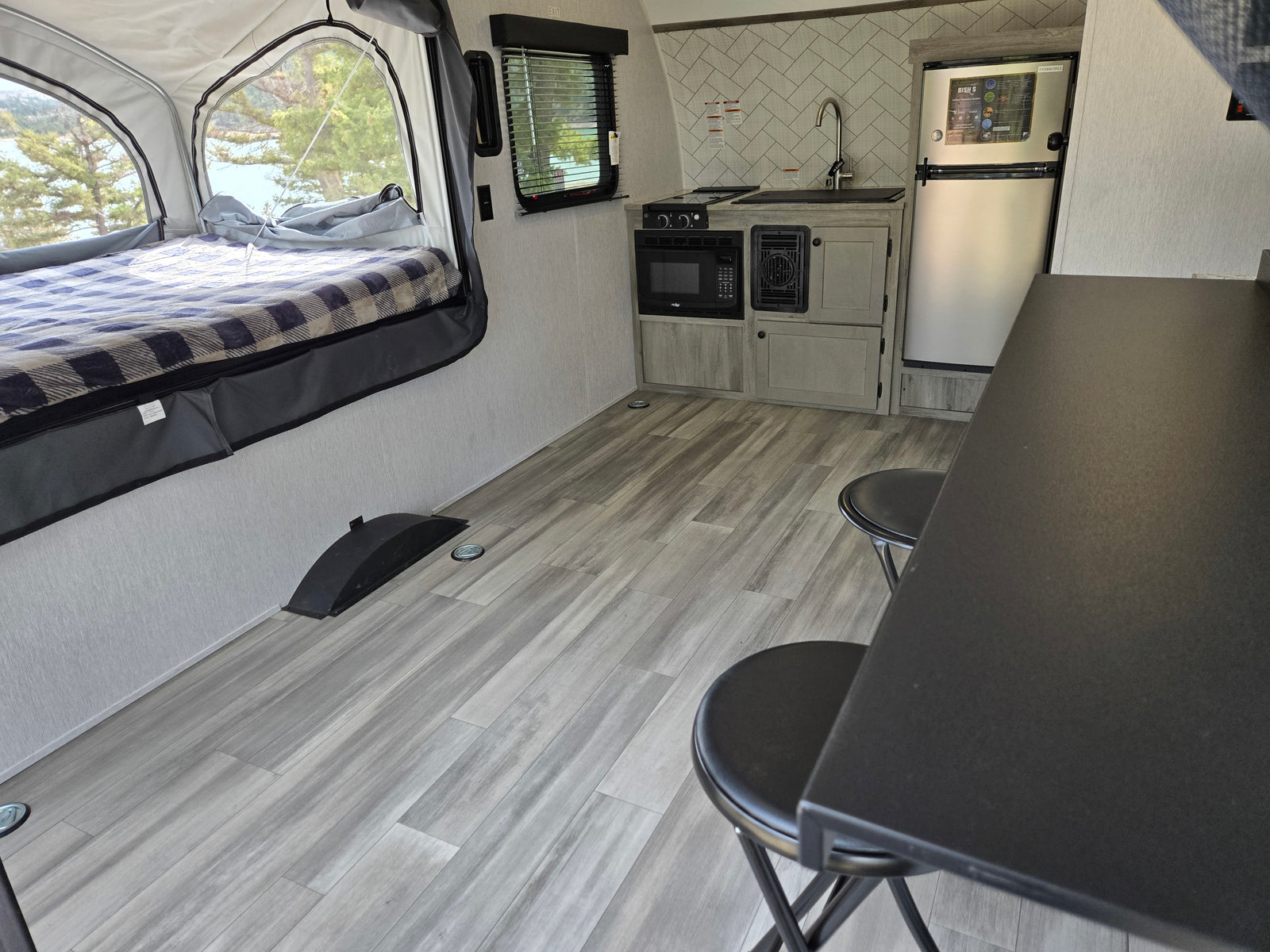 Cozy rv rental interior with pop-up bed, lake view, compact kitchen (microwave, stove, sink, fridge), and dinette with stools.