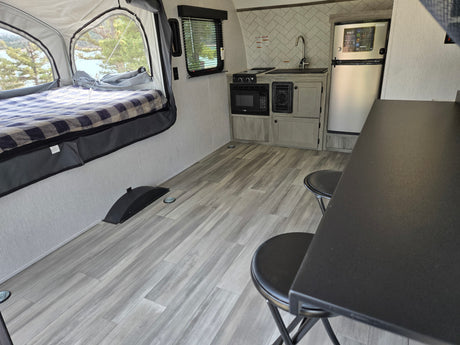 Cozy rv rental interior with pop-up bed, lake view, compact kitchen (microwave, stove, sink, fridge), and dinette with stools.