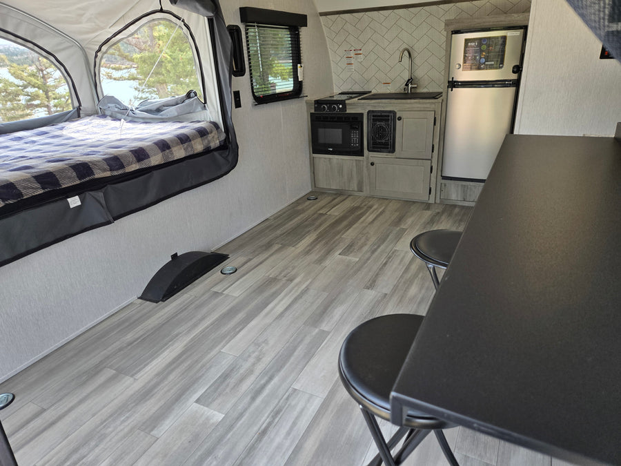 Cozy rv rental interior with pop-up bed, lake view, compact kitchen (microwave, stove, sink, fridge), and dinette with stools.