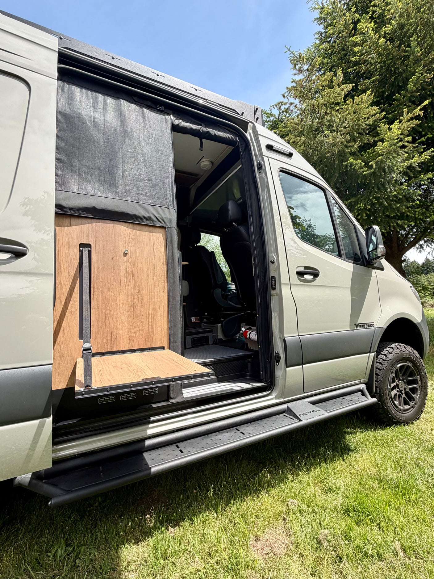 Experience an rv rental! A light green Winnebago Revel with an open side door, showing an exterior wooden table, rugged steps, & interior.