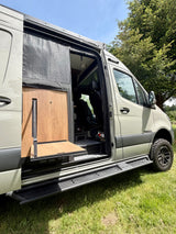 Experience an rv rental! A light green Winnebago Revel with an open side door, showing an exterior wooden table, rugged steps, & interior.