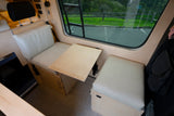 Inside an RV rental: A light wood interior with cream seating, an adjustable table, and bed area.
