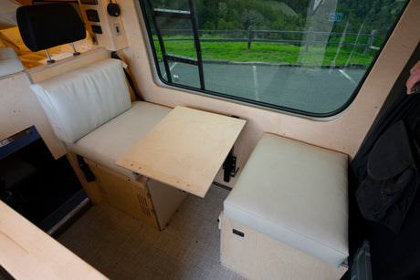Inside an RV rental: A light wood interior with cream seating, an adjustable table, and bed area.