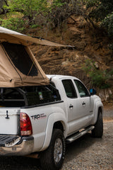 A white Toyota Tacoma pickup truck, equipped as an RV rental with a rooftop tent, parked on a gravel road, ready for off-road camping.