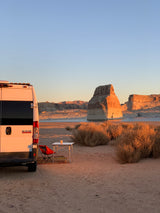 RV rental campsite at golden hour! A white camper van with a table & chair overlooks stunning desert rock formations & lake.