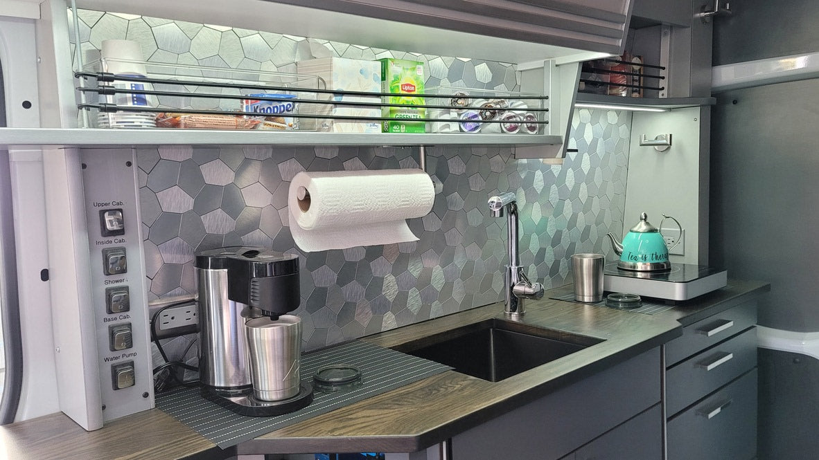 Well-equipped RV rental kitchen: sink, coffee maker, kettle, and overhead storage with Knoppers, Lipton tea, and K-cups.