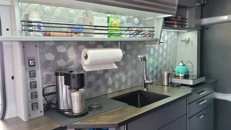 Well-equipped RV rental kitchen: sink, coffee maker, kettle, and overhead storage with Knoppers, Lipton tea, and K-cups.