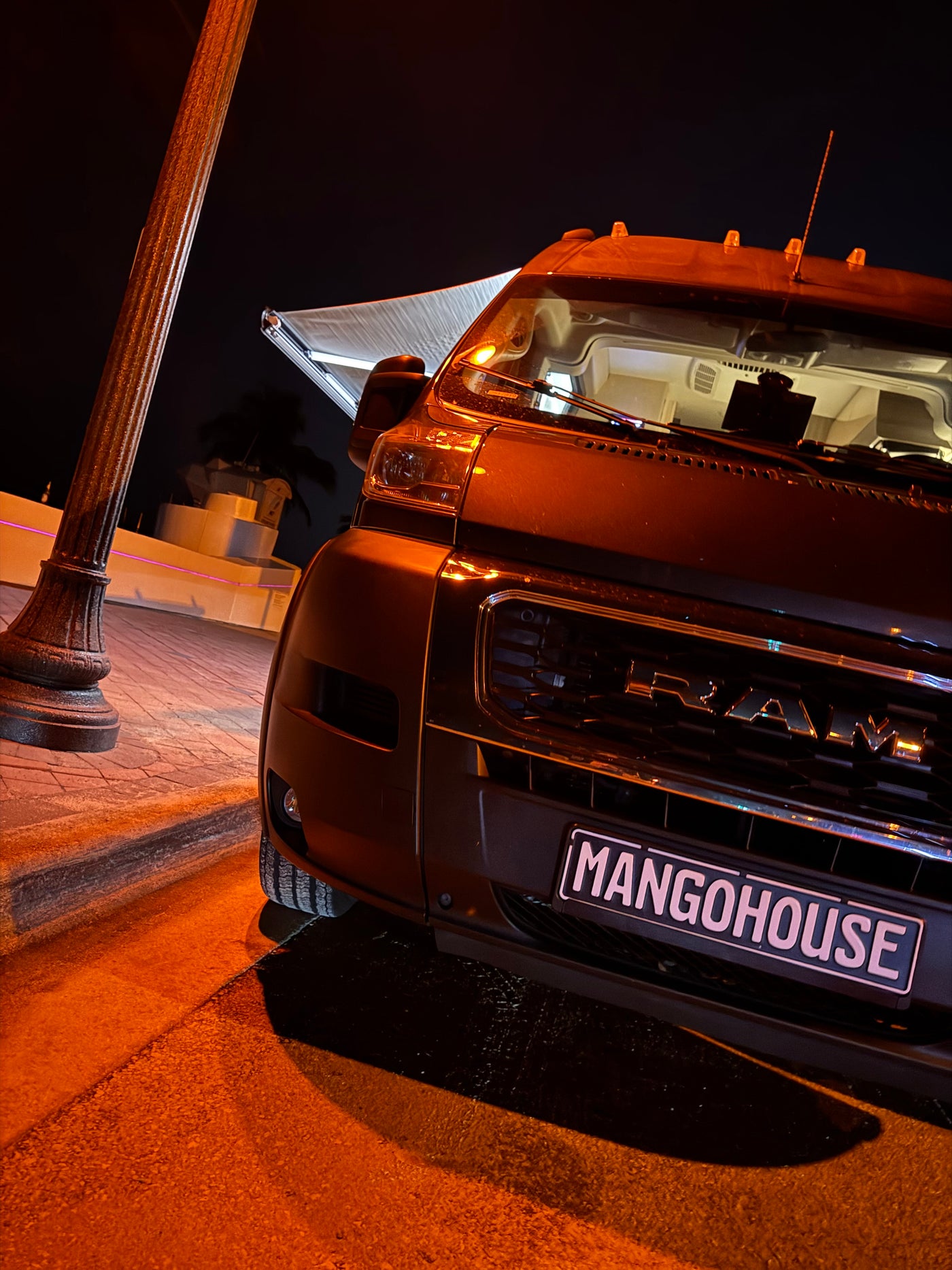 A sleek, dark MANGOHOUSE rv rental camper van with an extended awning, parked under warm, glowing lights at night. — photo 20