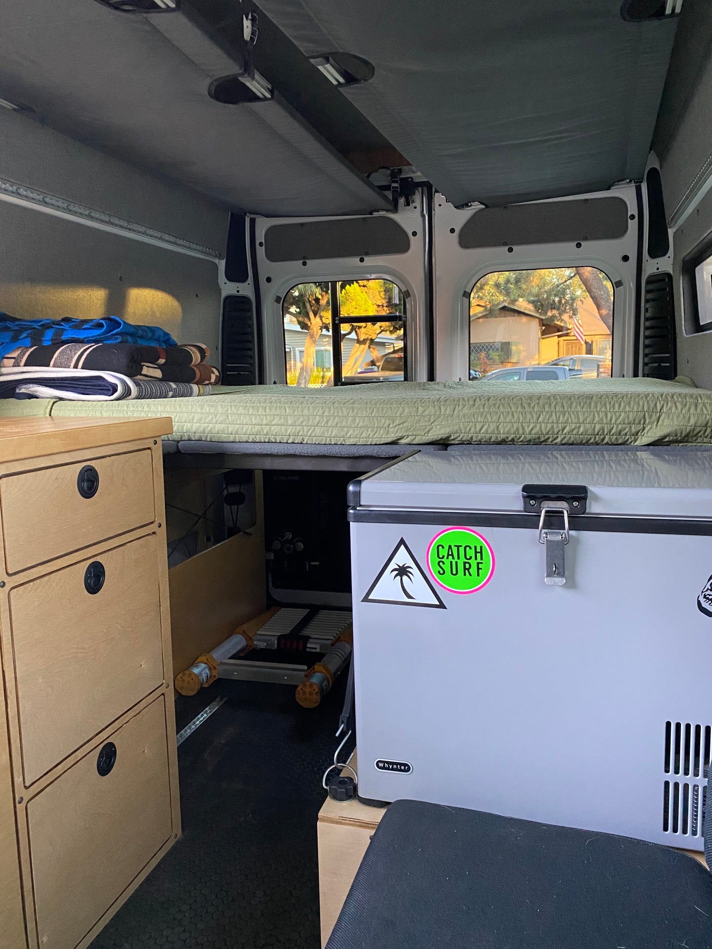 Inside this RV rental, a comfy bed rests above storage, alongside wooden drawers & a fridge, ideal for road trips.