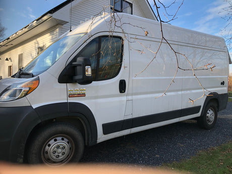 A white Ram ProMaster 2500 van, suitable for RV rental, is shown parked on a gravel driveway next to a white house.