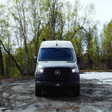 This grey Mercedes-Benz Sprinter RV rental is ready for adventure, on a muddy forest track with green trees and snow.