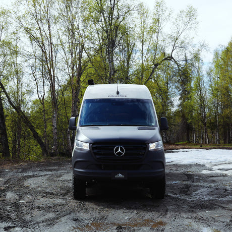 This grey Mercedes-Benz Sprinter RV rental is ready for adventure, on a muddy forest track with green trees and snow.