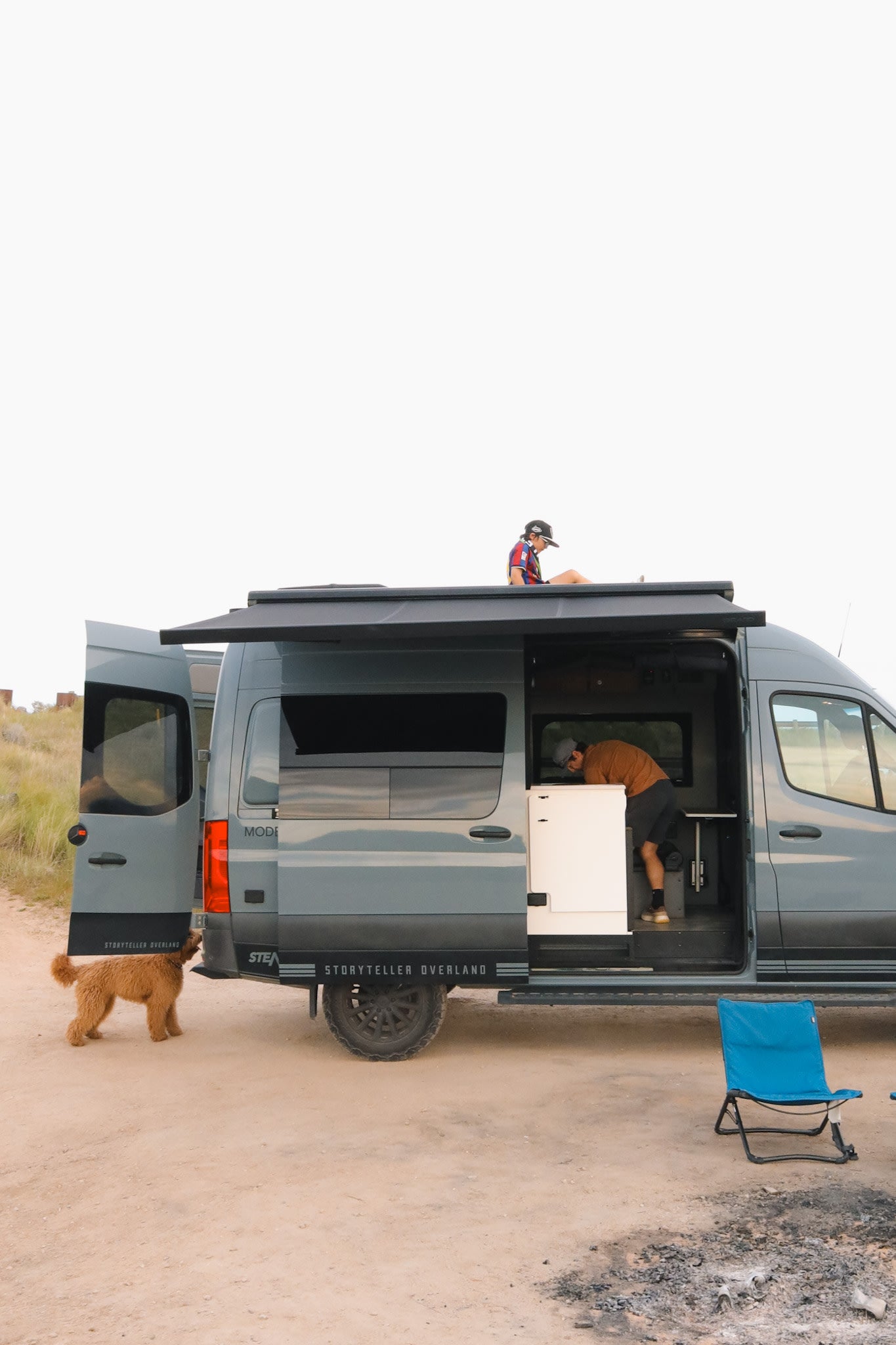 An open Storyteller Overland camper van, a perfect **RV rental** for adventure. People are on the roof & inside, with a dog exploring nearby.
