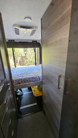 Inside an RV rental: rear bed, large window with forest view, AC, and under-bed storage. Ideal for travel.