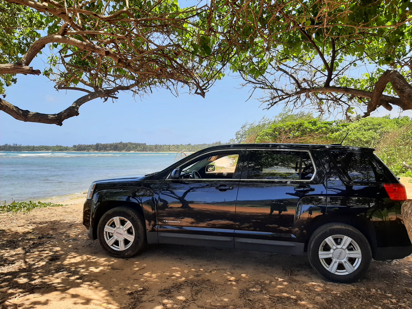 A black GMC Terrain SUV is parked under a shady tree by a beautiful beach. Perfect for coastal adventures, like those found with an RV rental.