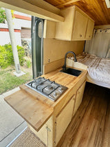 Compact RV rental kitchen & living space. Features a wood countertop with sink, 2-burner stove, pull-out table, & cozy bed area.
